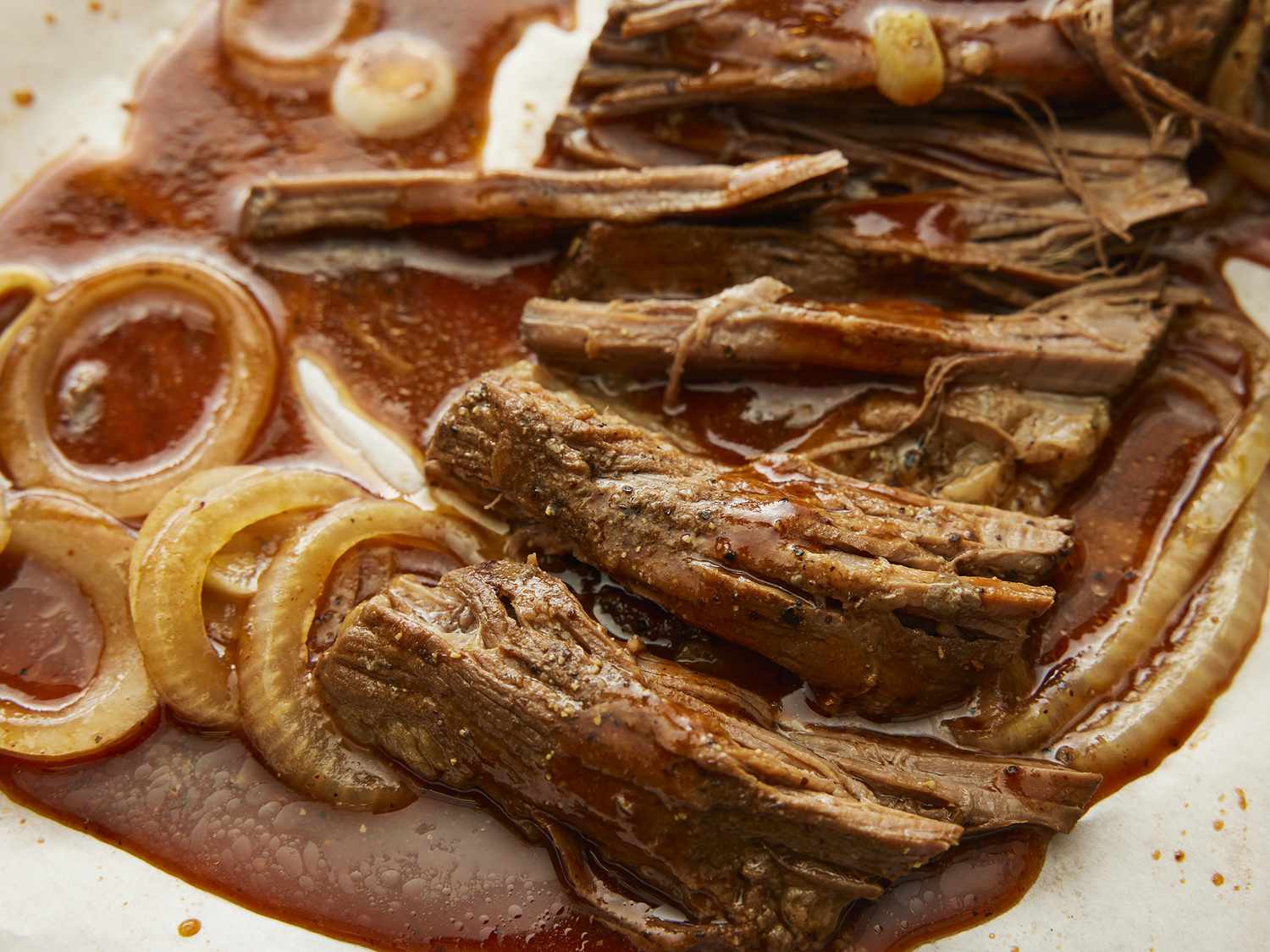 Raw beef brisket on a cutting board surrounded by sliced onion, garlic, beef broth, Worcestershire sauce, and spices on a kitchen counter