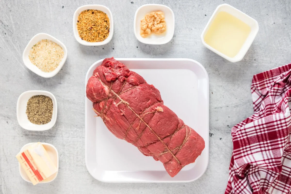 Raw beef tenderloin roast on a tray with small bowls of salt, pepper, chopped herbs, minced garlic, Dijon mustard, butter, olive oil, and a lemon on a clean kitchen counter