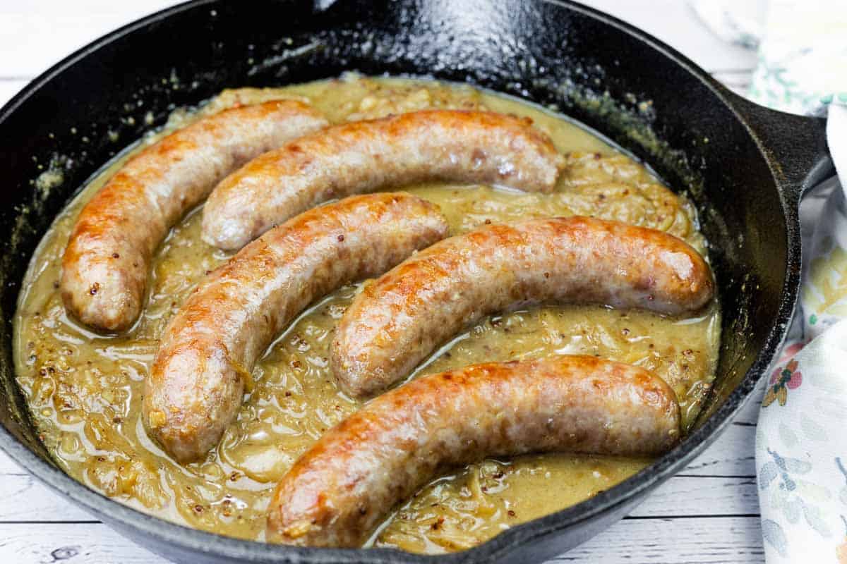 Raw bratwurst sausages, sliced onions, garlic, a bottle of beer, mustard, butter, and spices arranged on a light kitchen counter