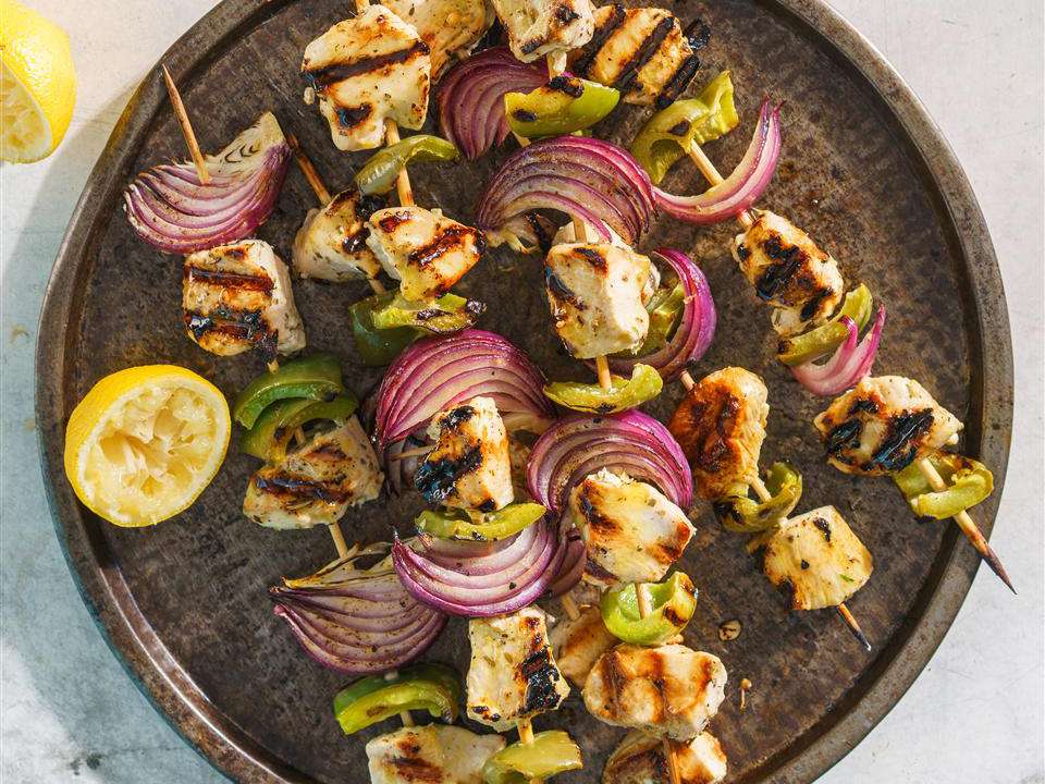 Raw chicken chunks, lemon, garlic, plain yogurt, olive oil, cumin, smoked paprika, salt, and black pepper arranged on a light kitchen counter with metal skewers nearby