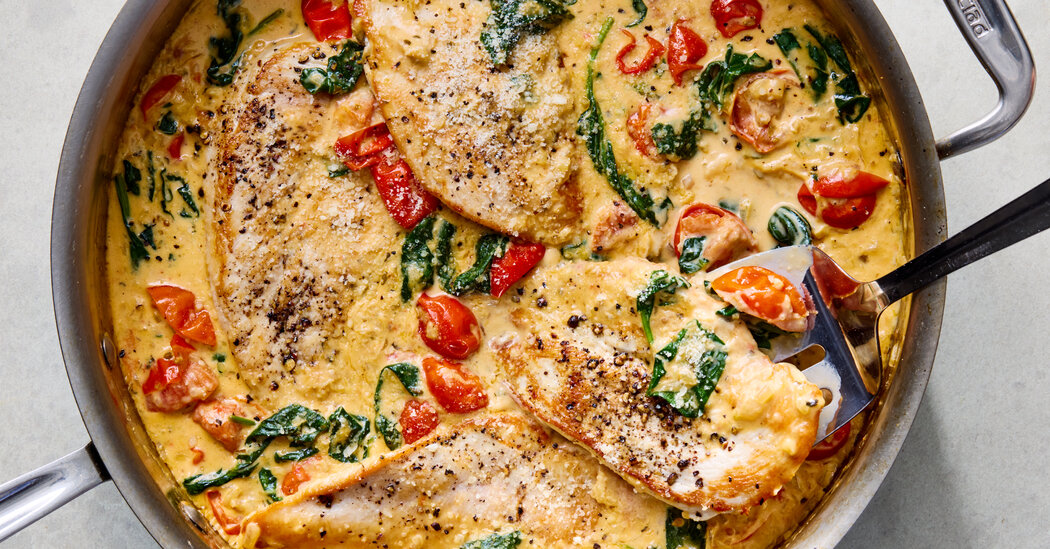 Raw chicken cutlets, garlic, baby spinach, sun-dried tomatoes, Parmesan, chicken broth, and cream arranged on a clean kitchen counter with a wooden cutting board