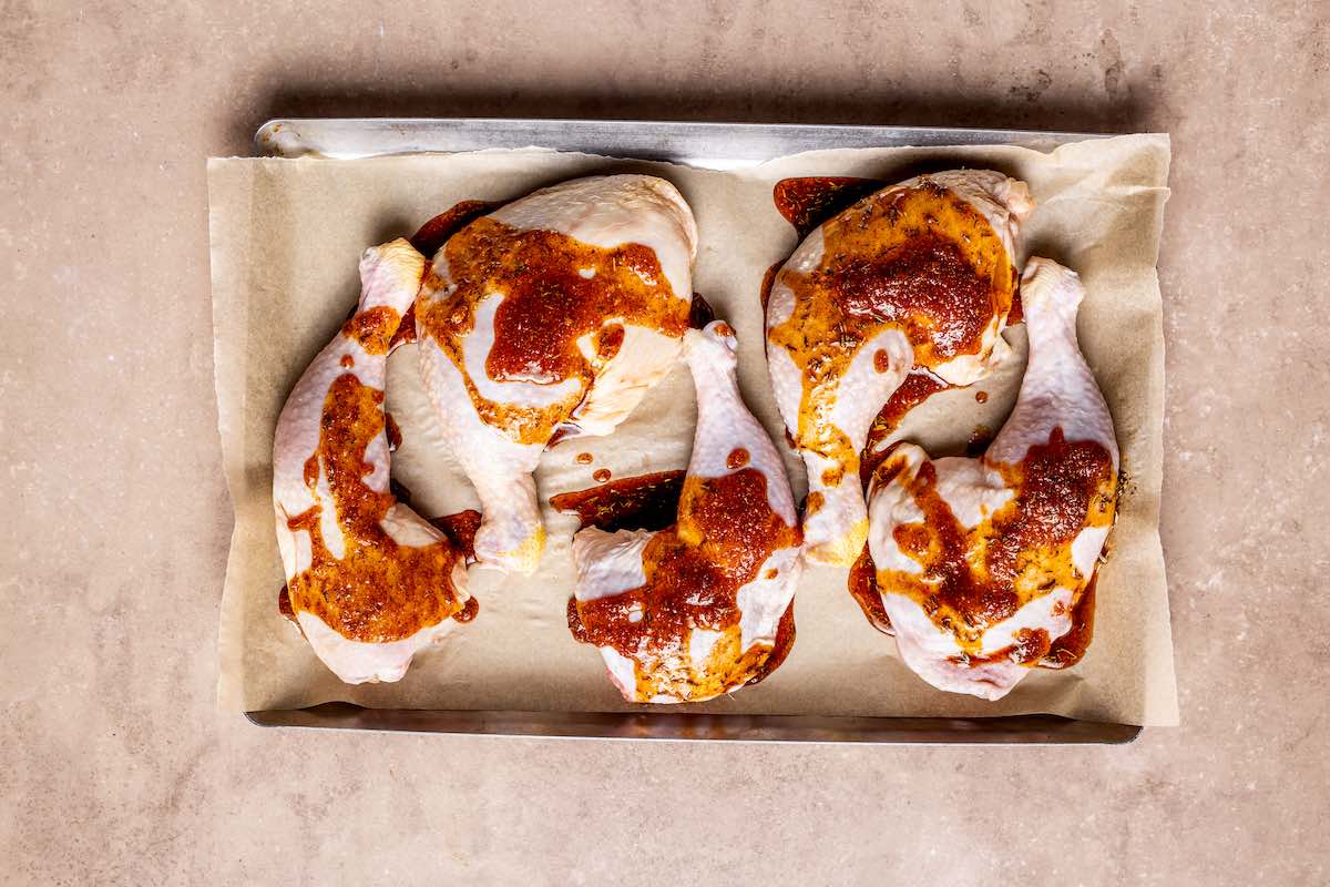 Raw chicken legs patted dry and coated in a reddish spice rub on a parchment-lined baking sheet
