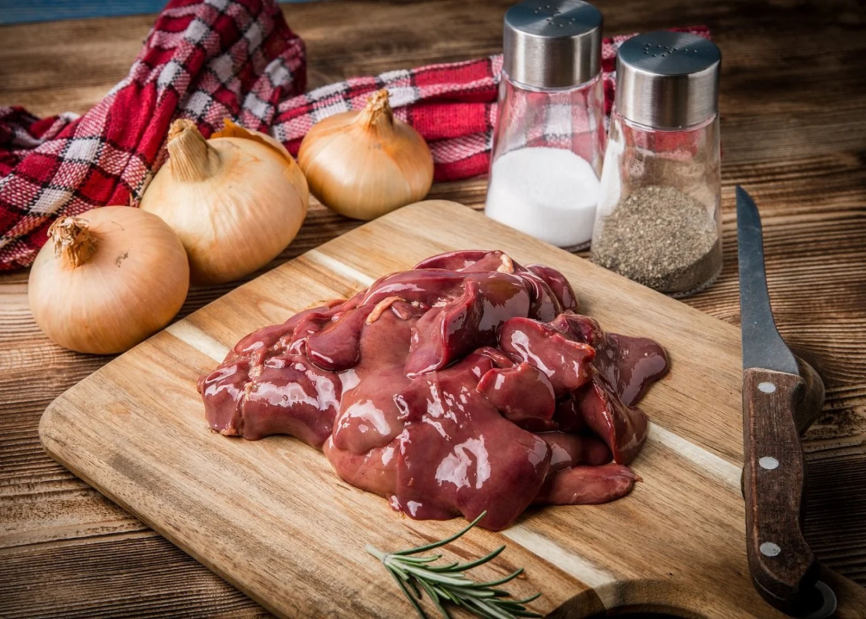 Raw chicken livers on a cutting board with small bowls of flour, capers, garlic, lemon, and butter arranged nearby on a kitchen counter
