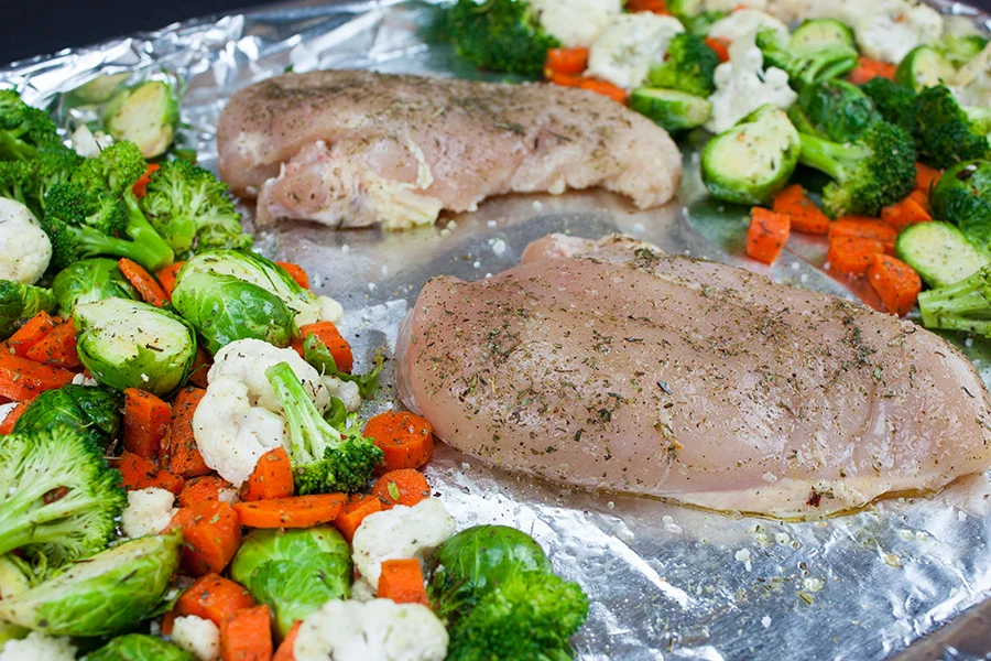 Raw chicken pieces and chopped broccoli, bell pepper, and red onion tossed with oil and spices on a parchment lined sheet pan before roasting