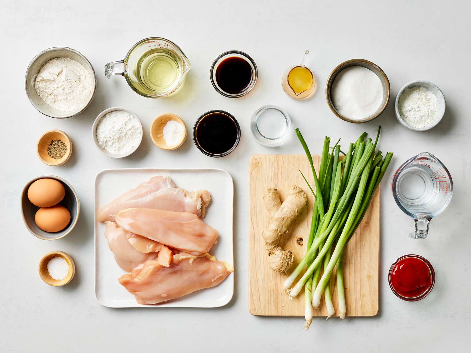 Raw chicken pieces, cornstarch, soy sauce, rice vinegar, hoisin, sugar, garlic, ginger, dried chiles, scallions, and sesame oil arranged on a bright kitchen counter