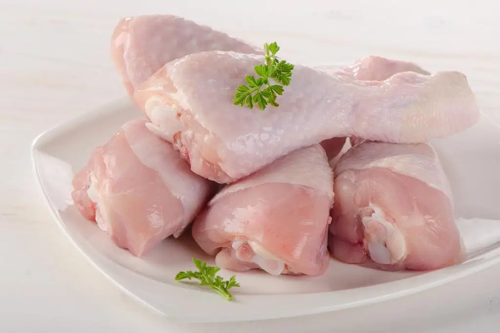 Raw chicken thighs and drumsticks on a cutting board beside smashed garlic cloves and bay leaves on a kitchen counter