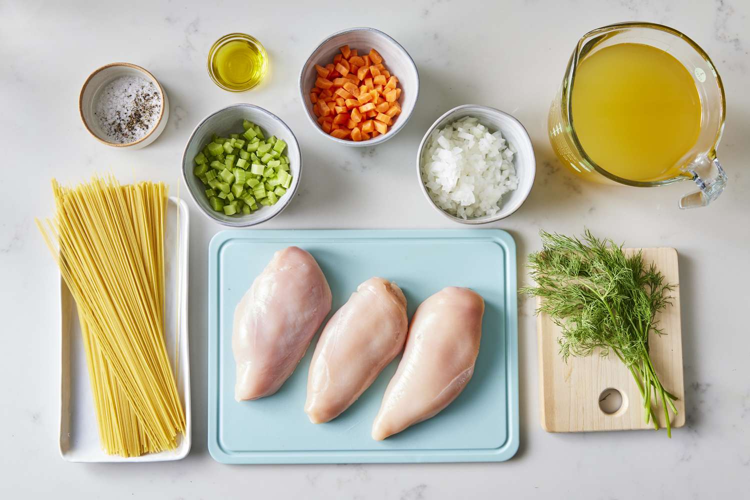 Raw chicken thighs, carrots, celery, onion, garlic, chicken broth, bay leaves, parsley, lemon, and egg noodles arranged on a clean kitchen counter in soft daylight
