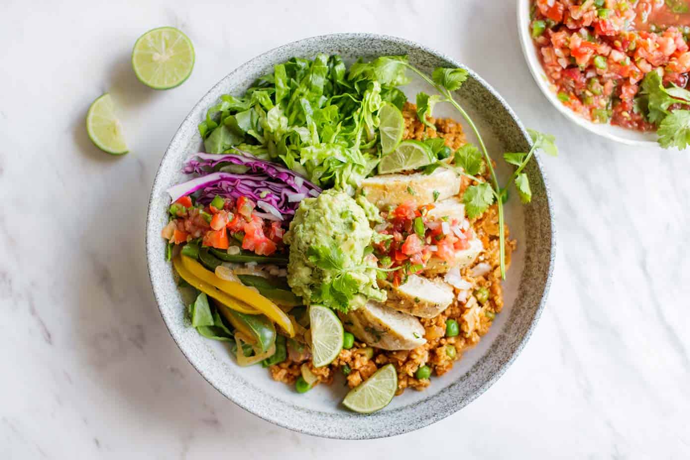 Raw chicken thighs, cauliflower rice, cucumber, cherry tomatoes, red onion, lemons, garlic, tahini, and small bowls of spices arranged on a bright kitchen counter