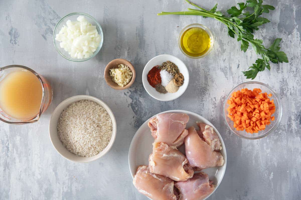 Raw chicken thighs, long grain rice, chicken broth, onions, garlic, lemon, and spices arranged on a tidy kitchen counter, overhead view in soft natural light