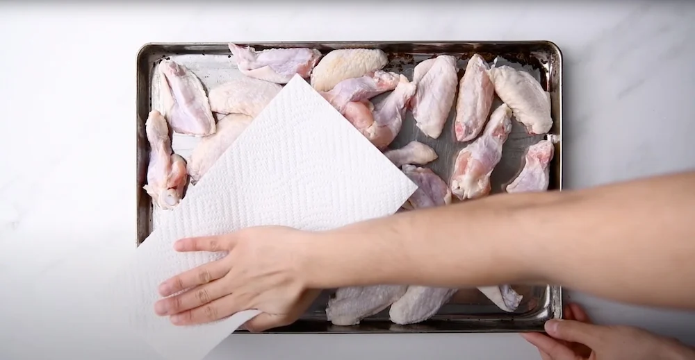 Raw chicken wings being patted dry with paper towels on a sheet pan