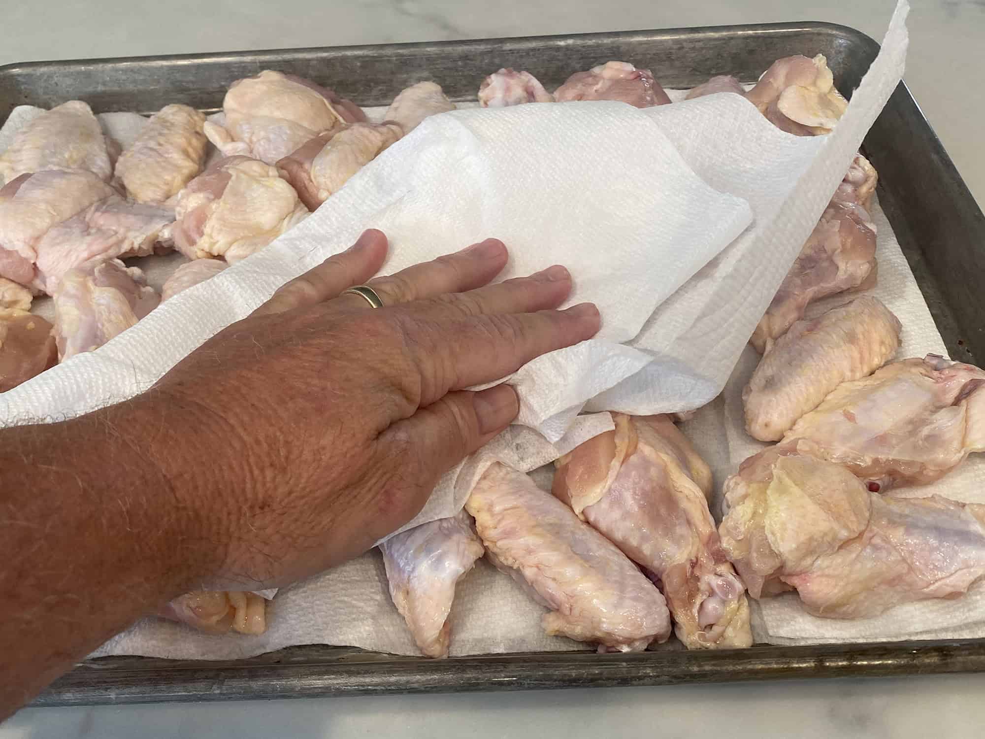 Raw chicken wings being patted very dry with paper towels on a sheet pan on a bright kitchen counter