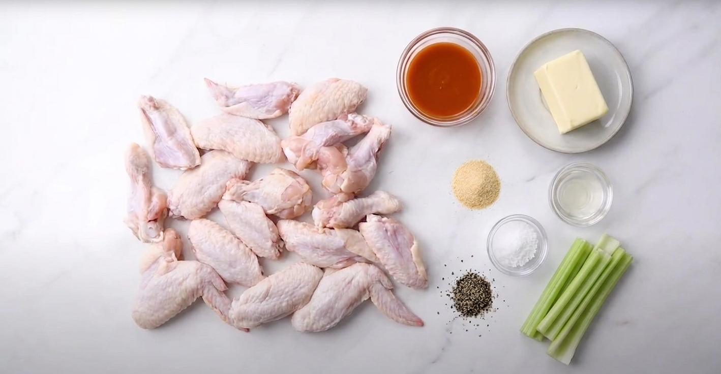 Raw chicken wings, hot sauce, butter, vinegar, garlic powder, salt, pepper, and celery arranged on a clean kitchen counter