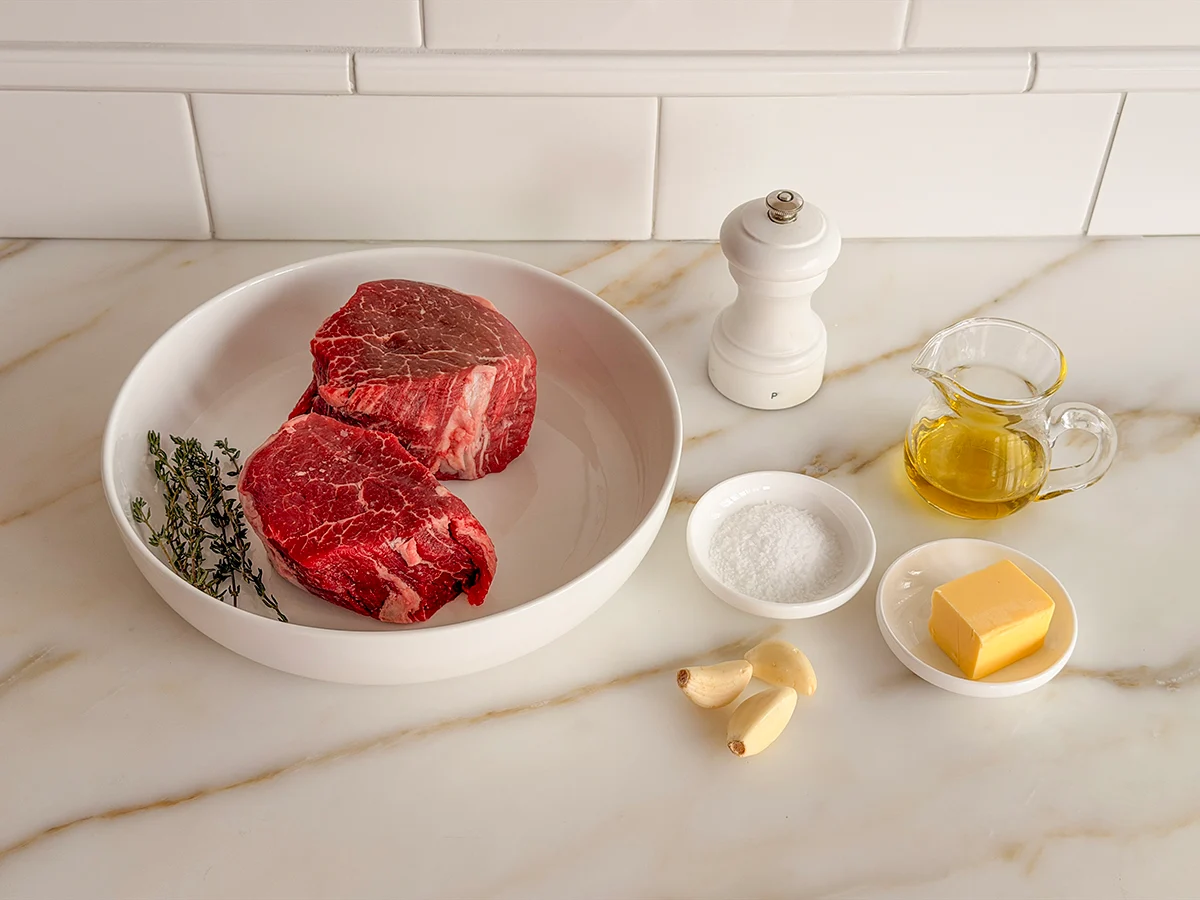 Raw filet mignon steaks on a cutting board with kosher salt, black pepper, garlic, butter, and fresh thyme arranged on a light kitchen counter