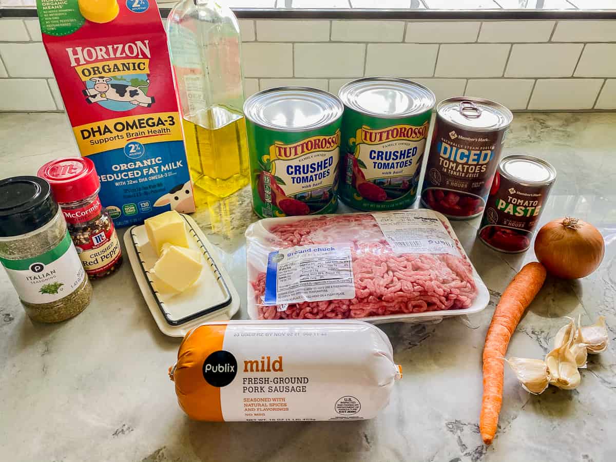 Raw ground beef, onions, carrots, celery, garlic, canned tomatoes, tomato paste, olive oil, red wine, and dried herbs arranged on a bright kitchen counter