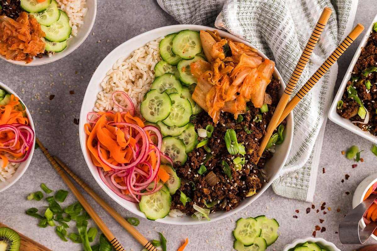 Raw ground beef, soy sauce, brown sugar, garlic, ginger, sesame oil, gochujang, rice, green onions, cucumber, and sesame seeds arranged on a clean kitchen counter