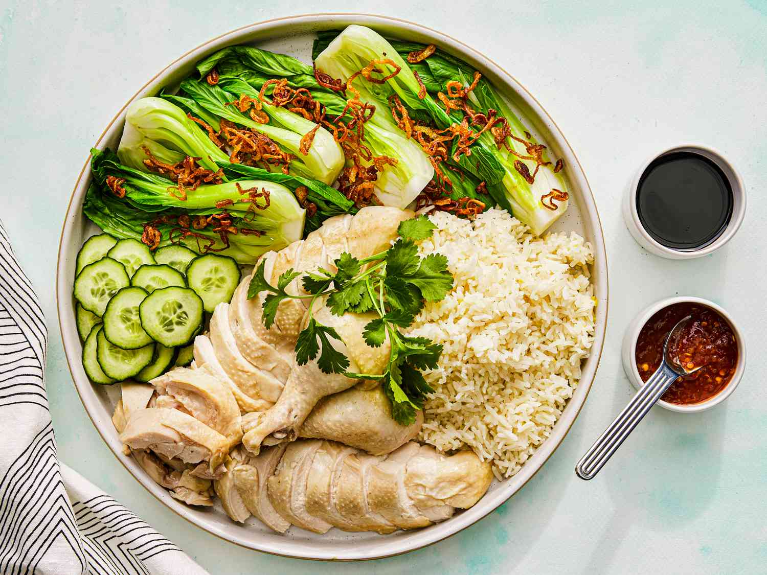 Raw ingredients for Hainanese chicken rice arranged on a kitchen counter: whole chicken, ginger, scallions, garlic, cucumber, jasmine rice, soy sauce, sesame oil, limes, and red chiles