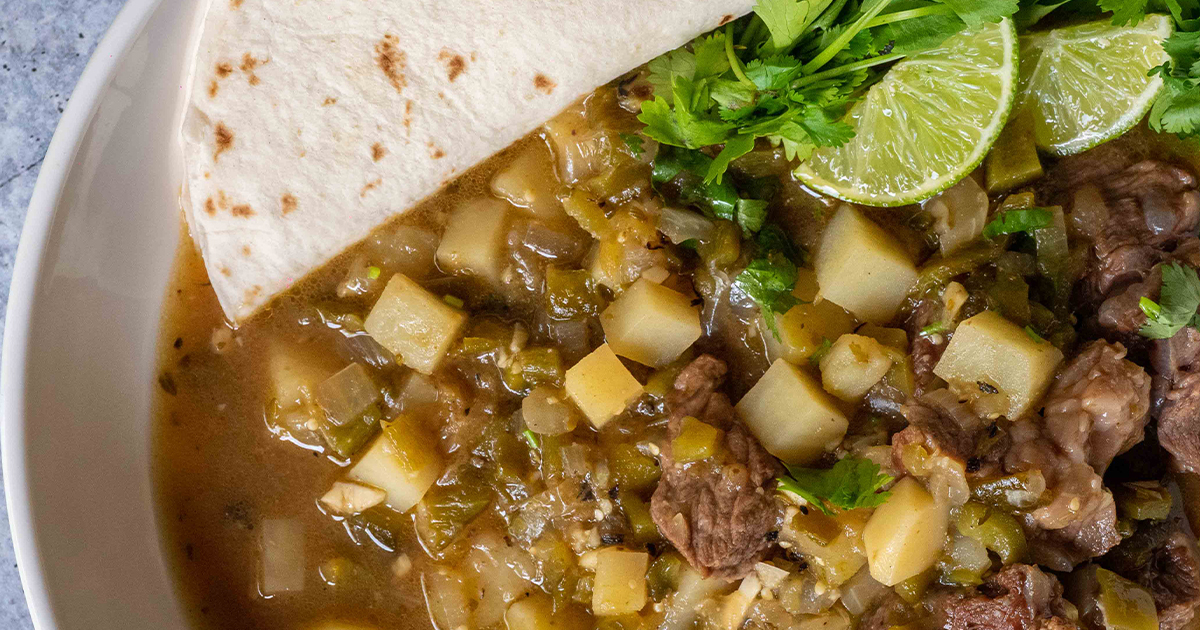 Raw ingredients for green chili pork stew arranged on a kitchen counter, including chopped onion, garlic, roasted green chiles, cubed pork, potatoes, and spices