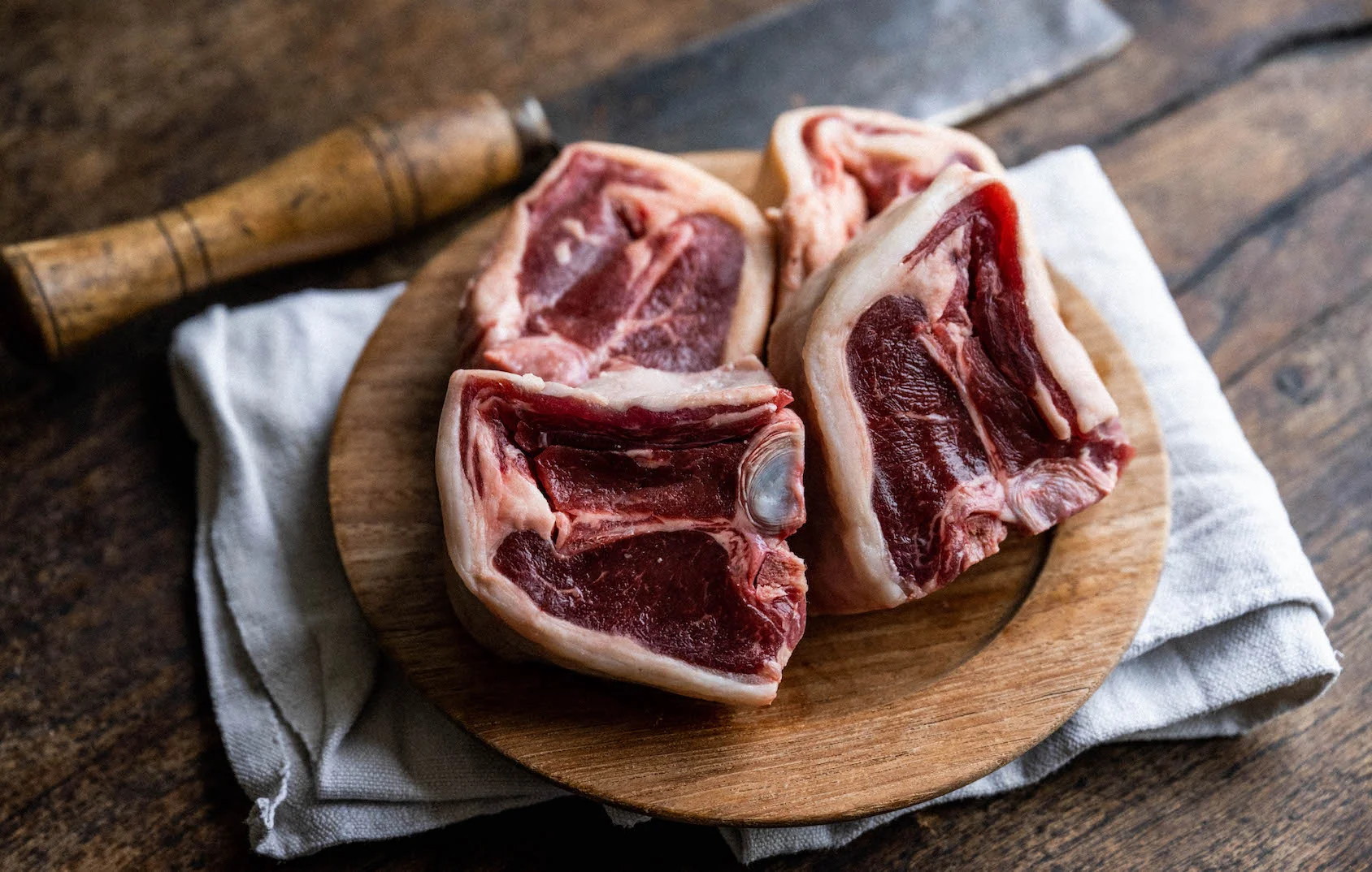 Raw lamb loin chops on a cutting board with small bowls of salt, pepper, chopped rosemary, minced garlic, lemon, and olive oil on a neutral kitchen counter