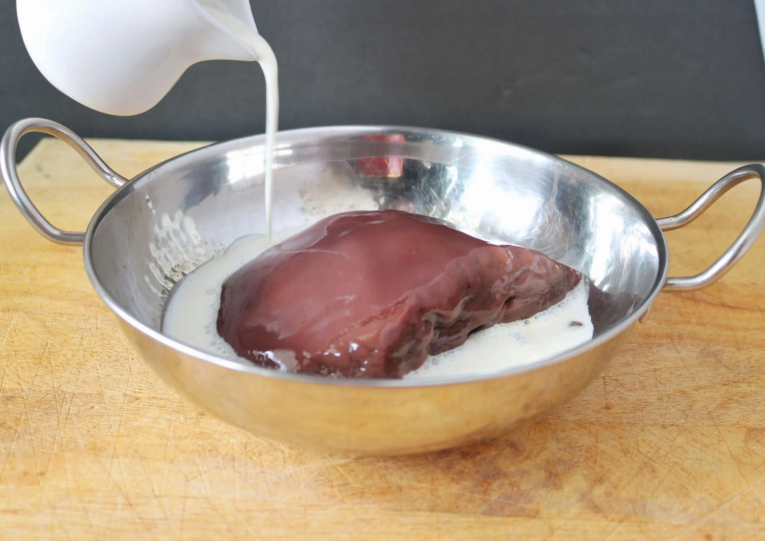 Raw liver slices submerged in milk in a shallow glass dish on a kitchen counter