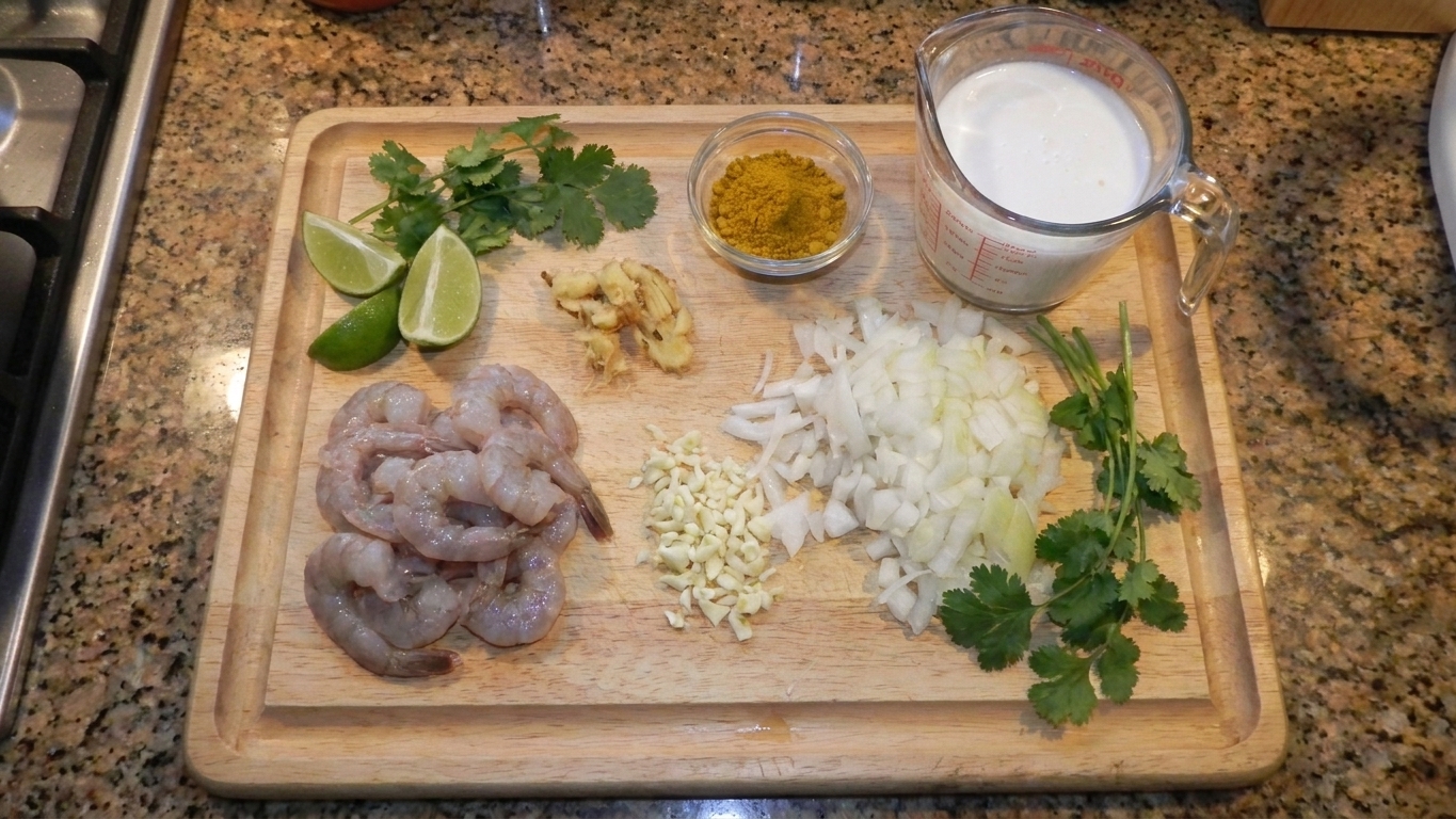 Raw peeled shrimp, coconut milk, curry powder, garlic, ginger, onion, lime, and cilantro arranged on a kitchen counter with a cutting board