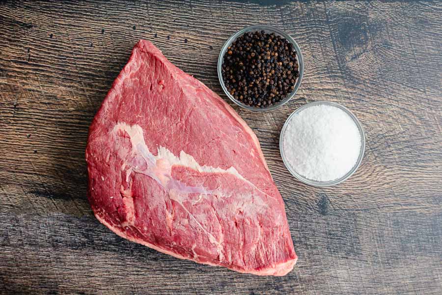 Raw picanha with a thick fat cap on a cutting board next to kosher salt, black pepper, garlic, butter, and a small bowl of chimichurri