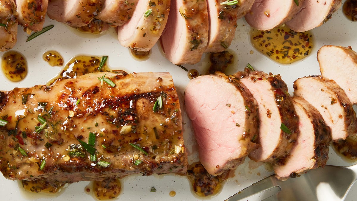 Raw pork fillet on a cutting board with small bowls of salt, pepper, garlic, herbs, lemon, mustard, and olive oil on a bright kitchen counter