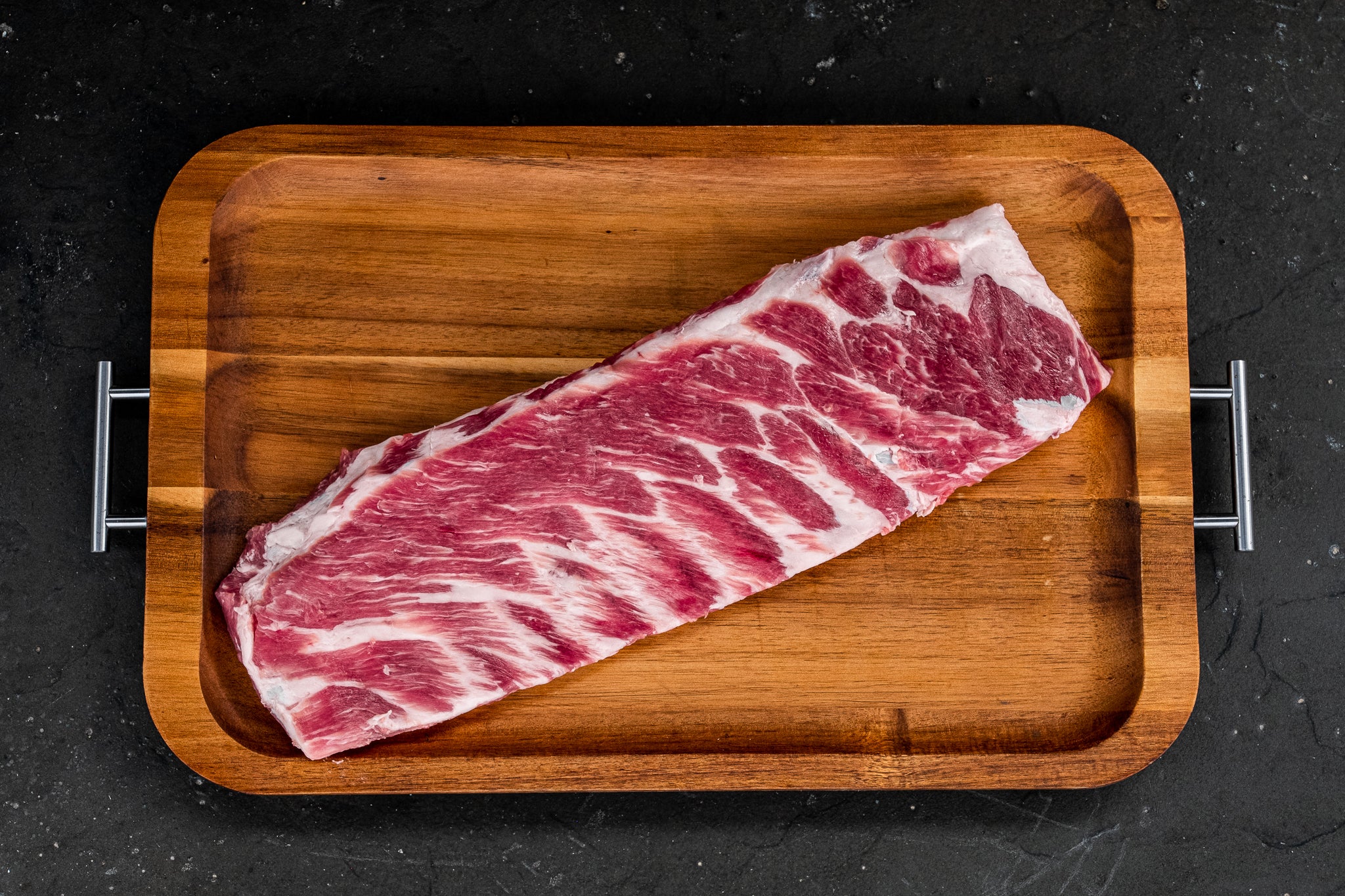Raw pork ribs on a cutting board surrounded by small bowls of brown sugar, paprika, garlic powder, salt, pepper, and a bottle of barbecue sauce on a kitchen counter