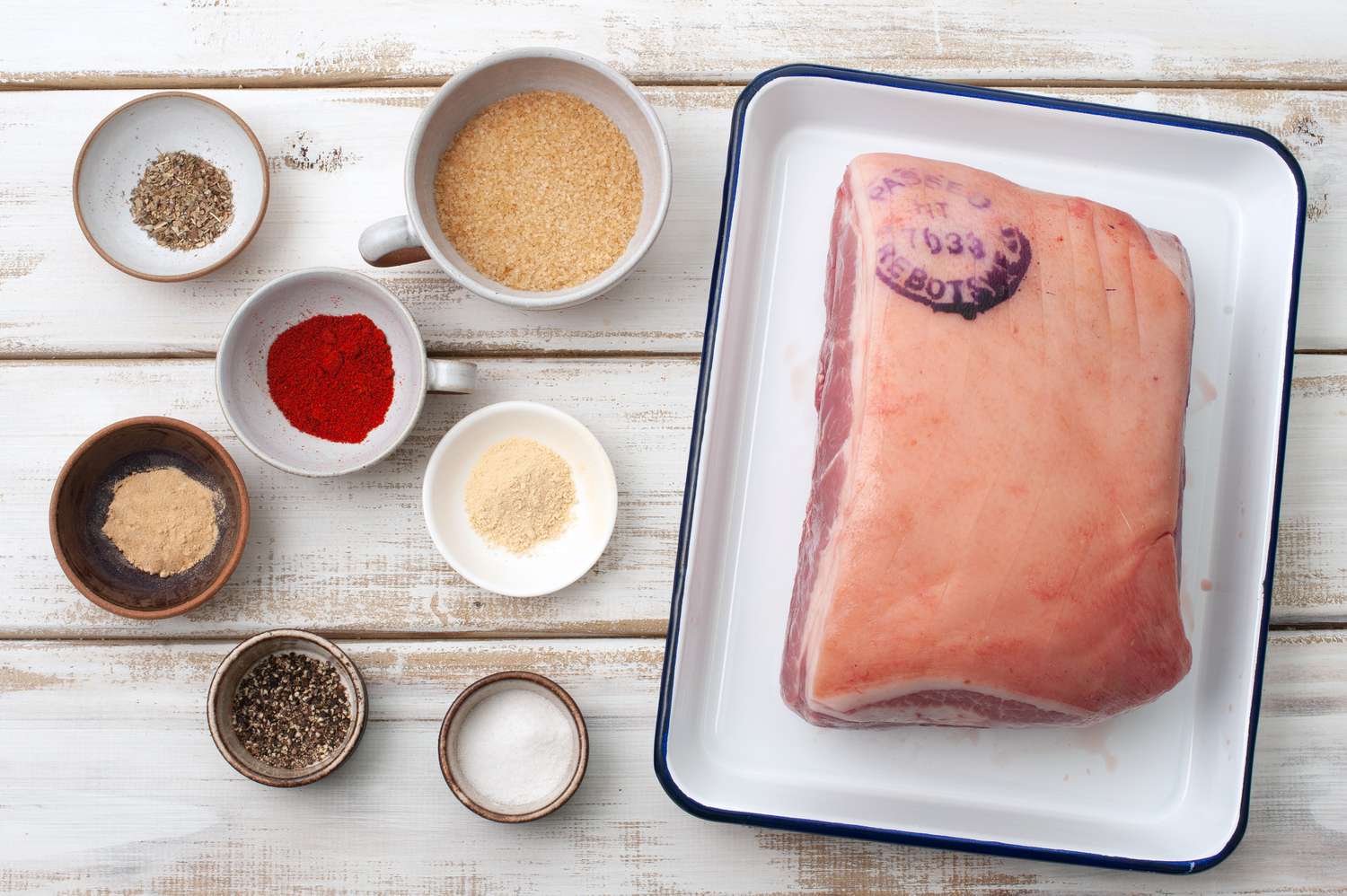 Raw pork shoulder on a cutting board surrounded by small bowls of salt, cumin, oregano, smoked paprika, minced garlic, and lime