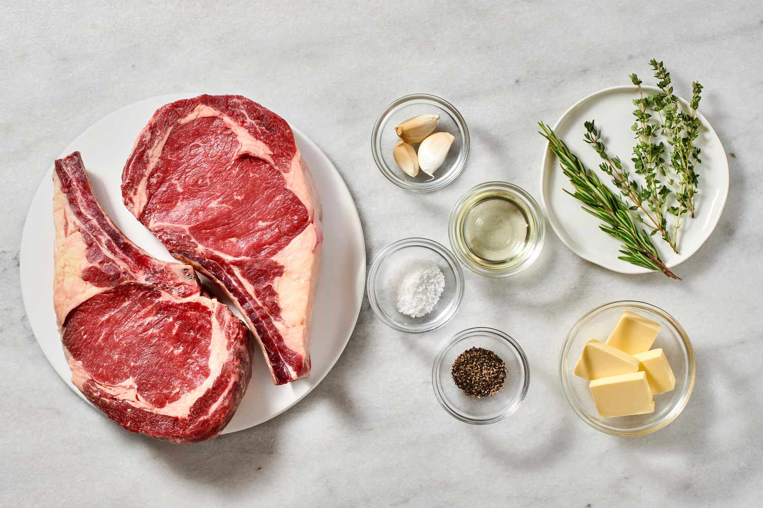 Raw ribeye steaks on a sheet pan next to kosher salt, black pepper, garlic cloves, fresh rosemary, fresh thyme, and a stick of butter on a kitchen counter