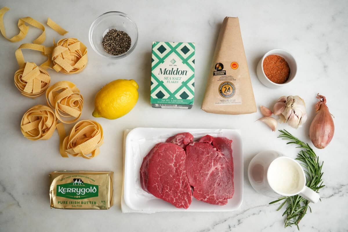 Raw ribeye steaks, uncooked pasta, garlic, lemon, Parmesan, parsley, and butter arranged on a light kitchen counter with a cutting board