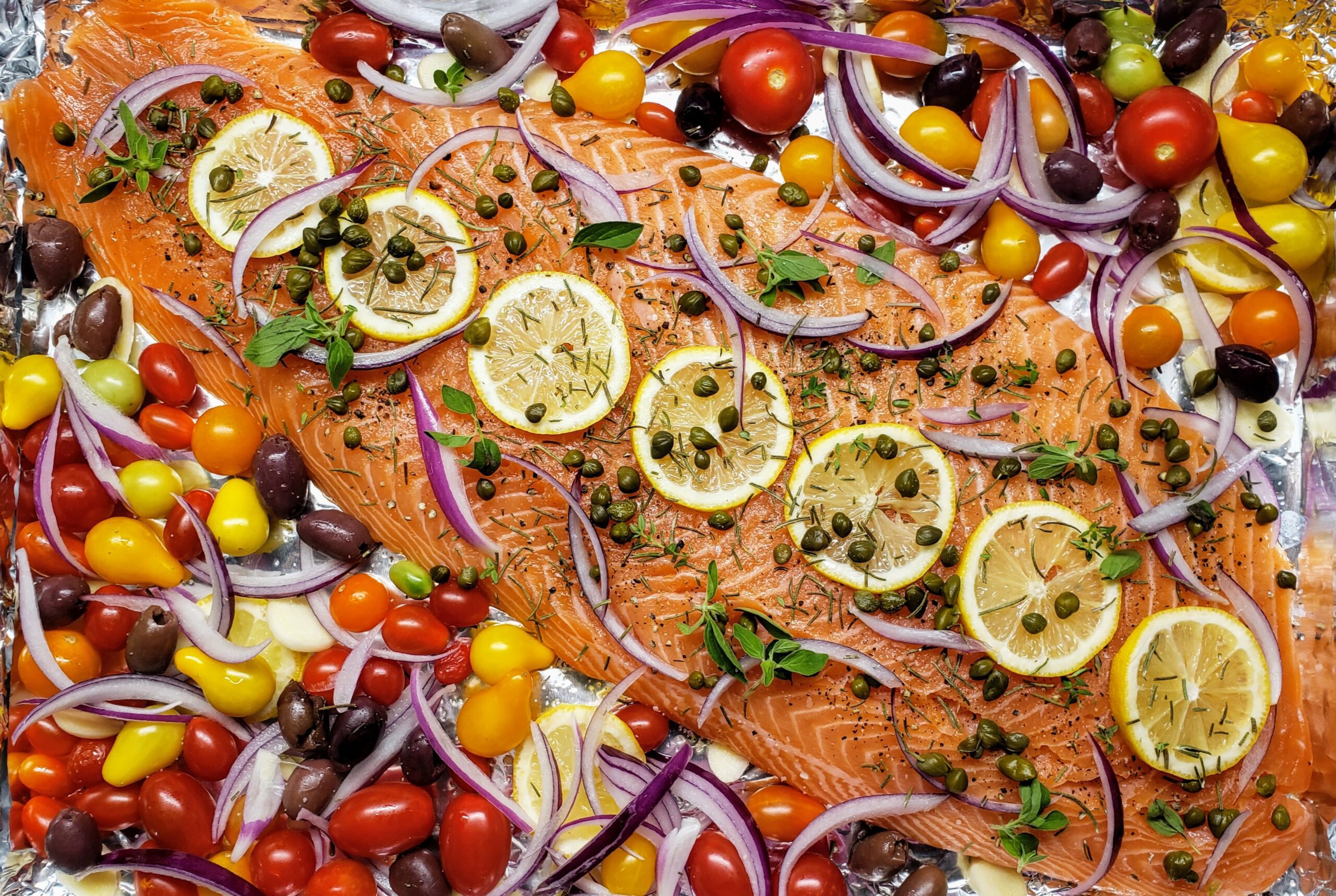 Raw salmon fillets, cherry tomatoes, Kalamata olives, feta, lemons, garlic, olive oil, and fresh parsley arranged on a bright kitchen counter with a cutting board