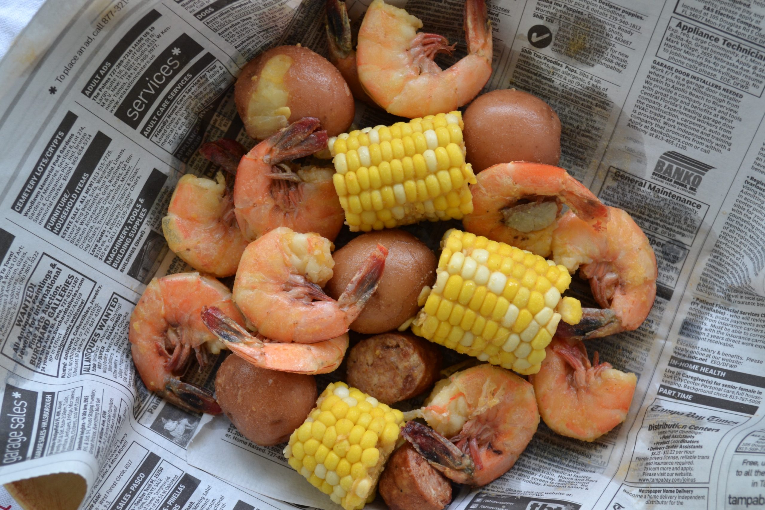 Raw shrimp, smoked sausage, corn, baby potatoes, lemons, garlic, and bowls of spices arranged on a light kitchen counter