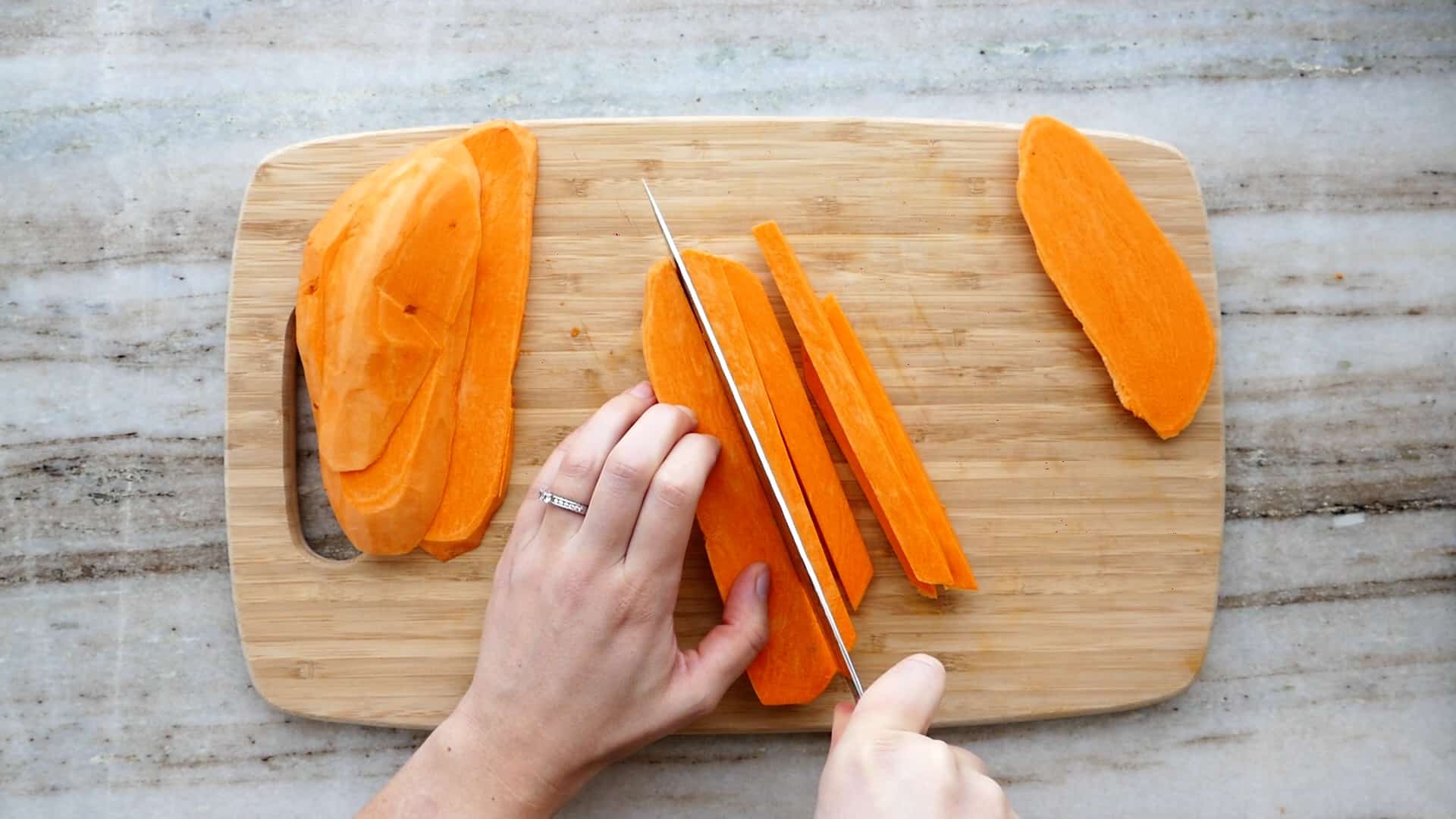 Raw sweet potato fries cut into evenly sized sticks on a wooden cutting board beside a chef's knife