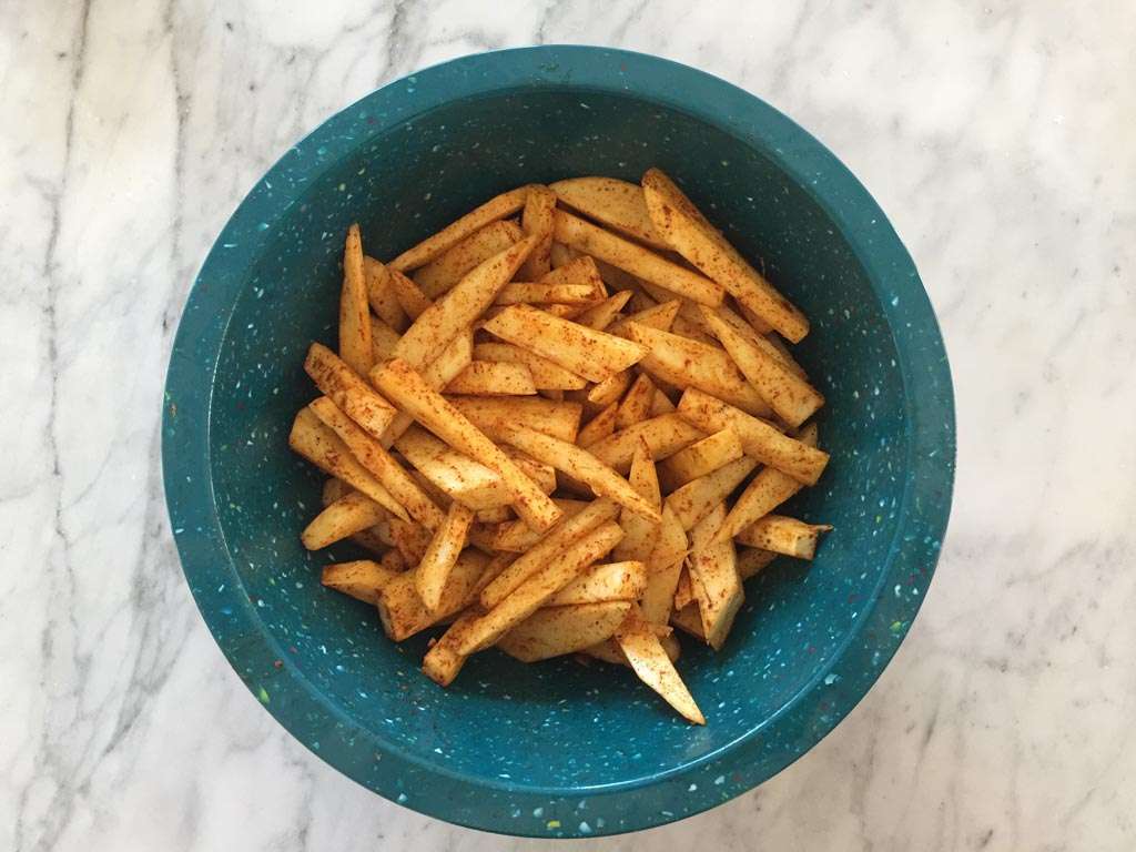Raw sweet potato fries in a mixing bowl coated in cornstarch and spices, ready to be air fried on a kitchen counter