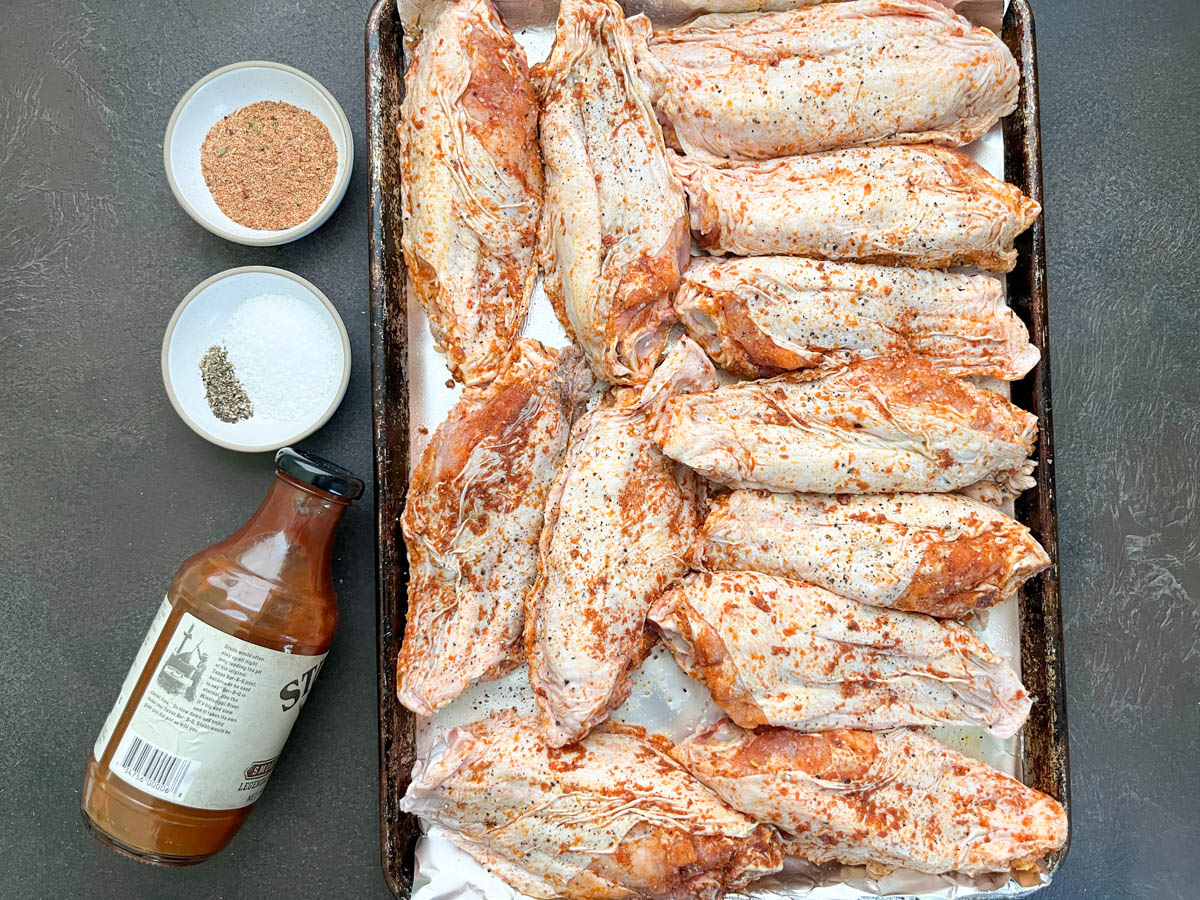 Raw turkey wings on a rimmed baking sheet with small bowls of spices, dried herbs, and oil on a kitchen counter