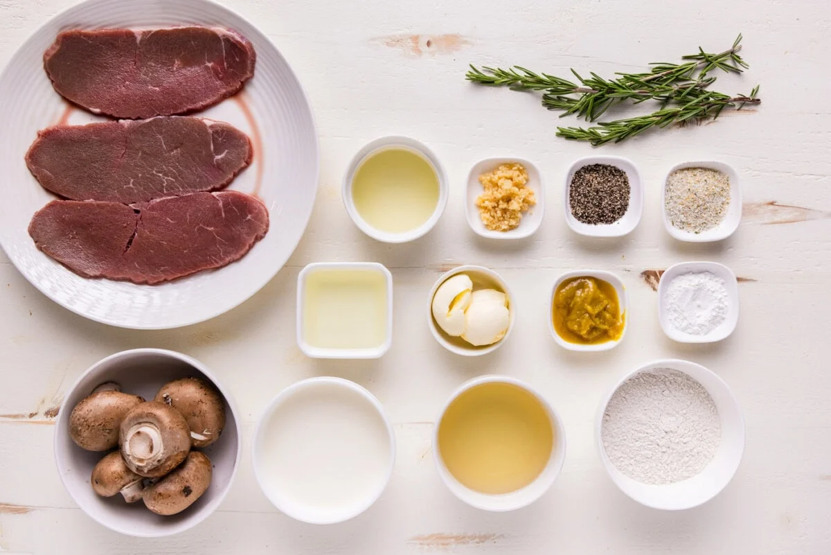 Raw veal cutlets, lemons, capers, garlic, flour, butter, parsley, and a small bowl of chicken broth arranged on a tidy kitchen counter in warm natural light