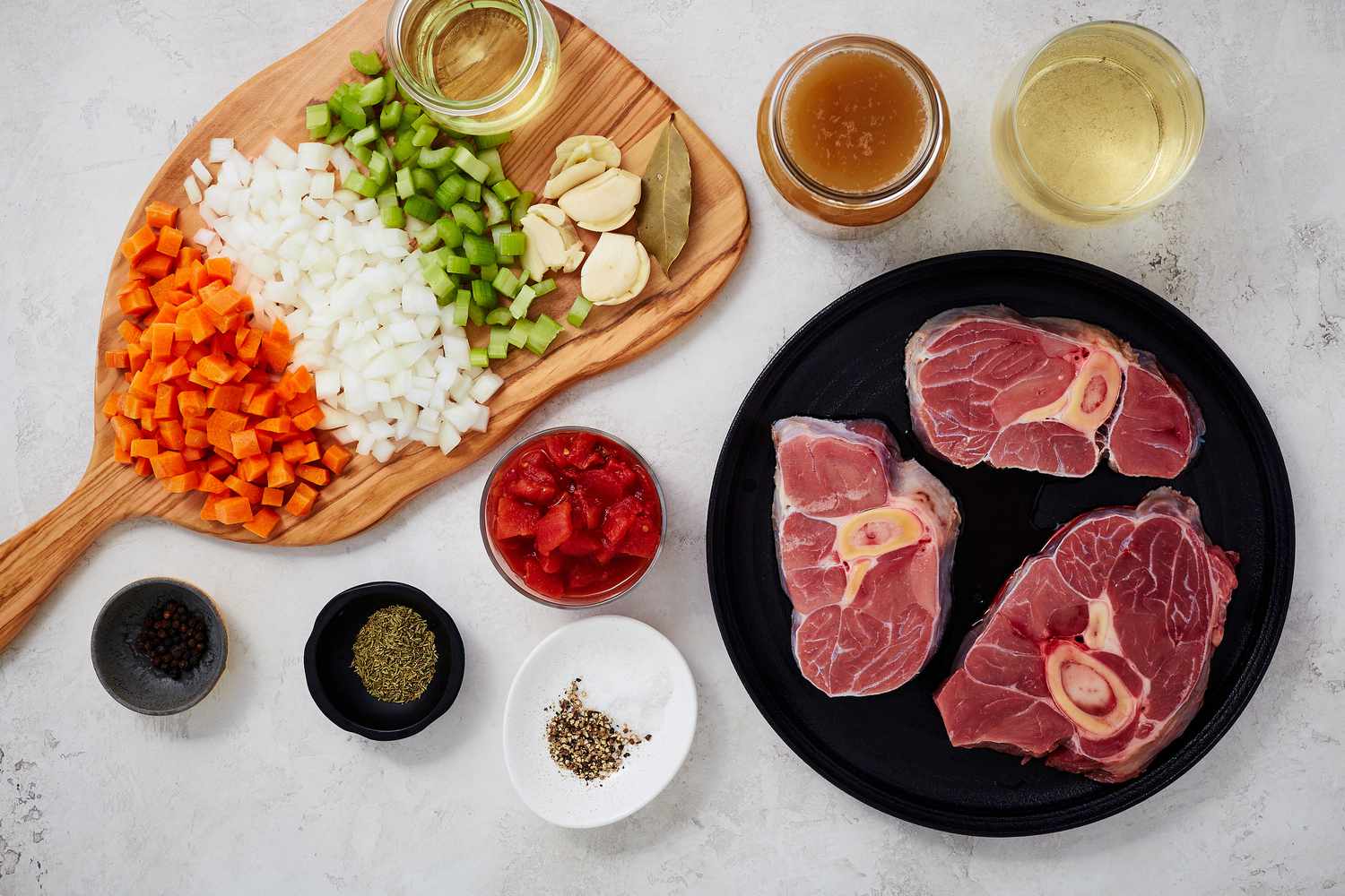 Raw veal or beef shanks, carrots, celery, onion, garlic, tomato paste, canned tomatoes, wine, broth, and fresh parsley arranged on a clean kitchen counter in warm natural light