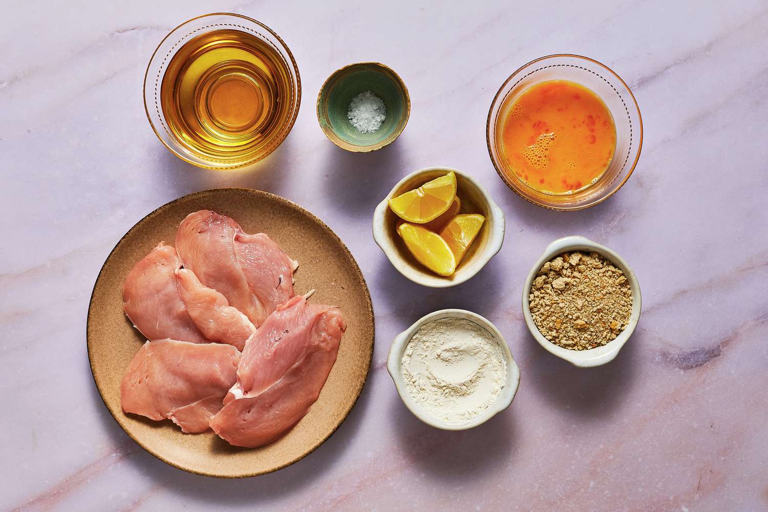 Raw veal or pork cutlets, eggs, flour, breadcrumbs, lemon, parsley, and a shallow dish of oil arranged on a clean kitchen counter in consistent warm lighting
