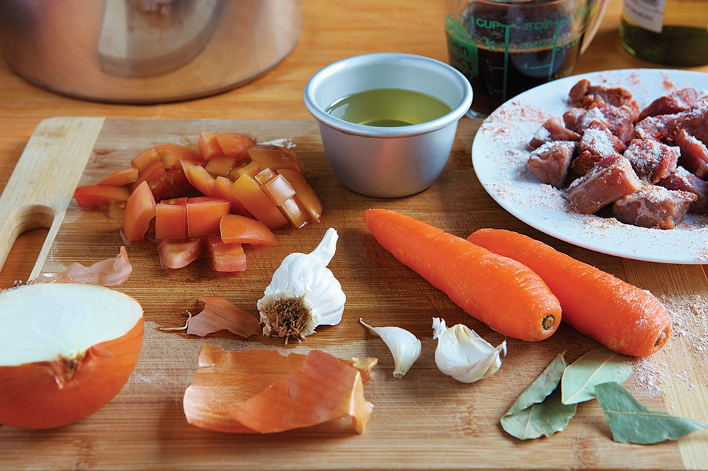Raw veal shanks on a cutting board with onions, carrots, celery, garlic, canned tomatoes, white wine, and fresh parsley arranged on a kitchen counter