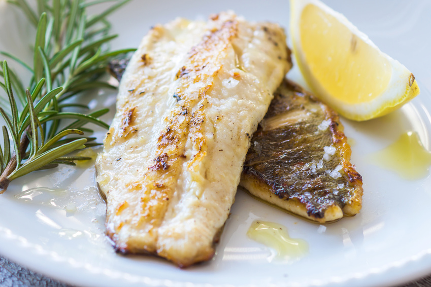 Raw white fish fillets, lemons, garlic, butter, parsley, olive oil, and spices arranged on a light kitchen counter with a cutting board