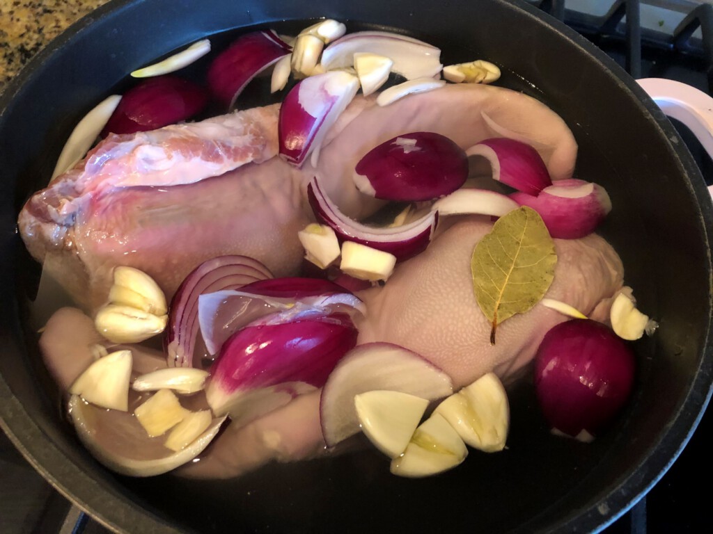 Raw whole beef tongue on a cutting board next to onion, garlic, bay leaves, peppercorns, and limes on a clean kitchen counter