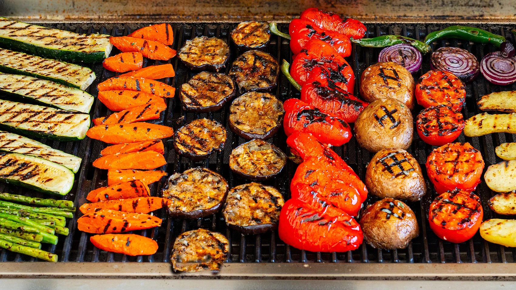 Raw zucchini planks, bell pepper strips, red onion wedges, mushrooms, and asparagus arranged on a kitchen counter with small bowls of olive oil, salt, pepper, lemon, and chopped herbs