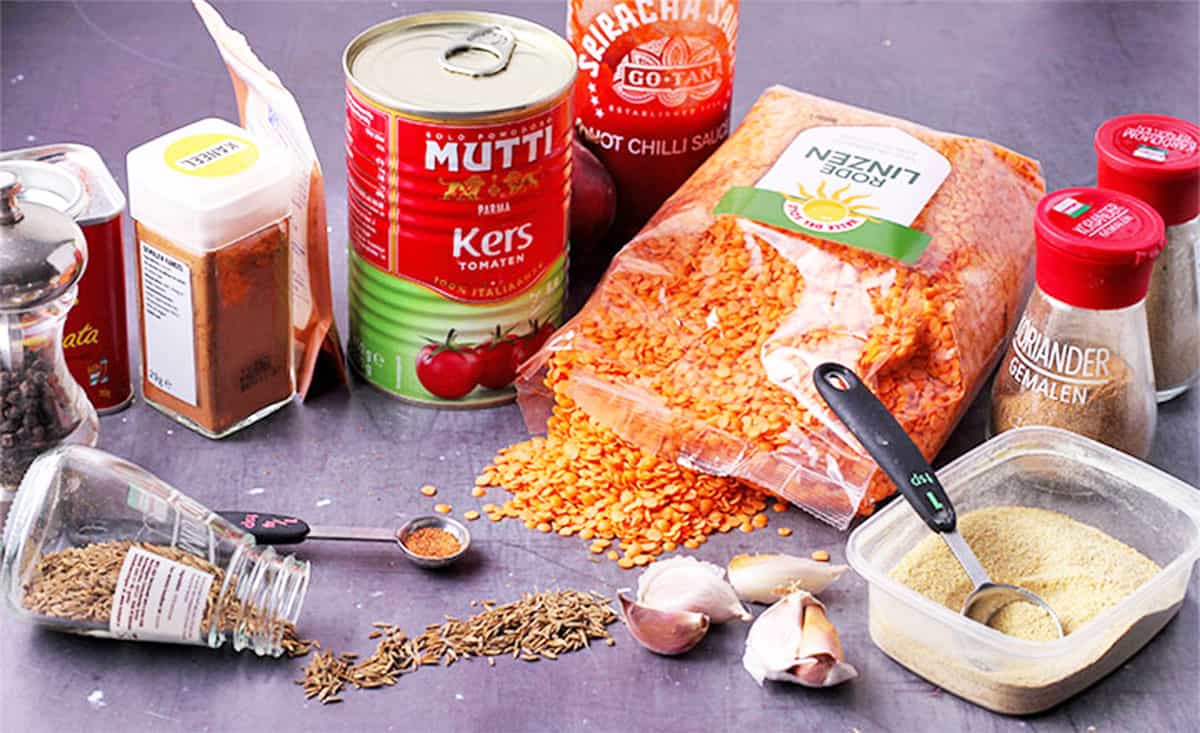 Red lentils in a small bowl surrounded by cumin seeds, turmeric, garam masala, onions, garlic, ginger, and canned tomatoes arranged on a kitchen counter