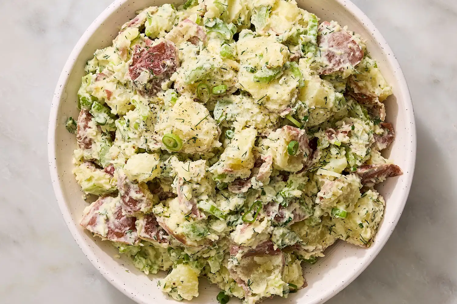 Red potatoes, mayonnaise, Dijon mustard, celery, red onion, fresh dill, parsley, lemon, and seasonings arranged on a light kitchen counter in a home kitchen