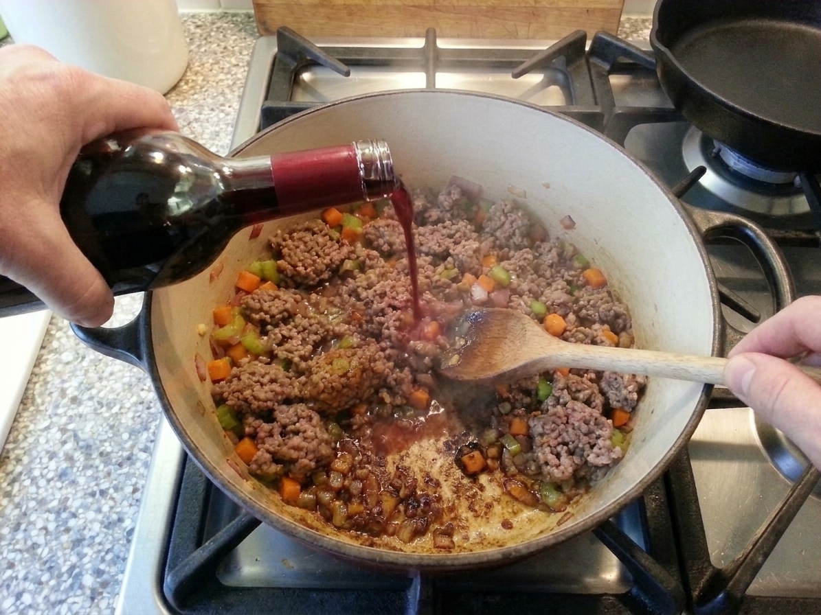 Red wine being poured into a pot of browned beef and vegetables as the cook scrapes the bottom of the pot