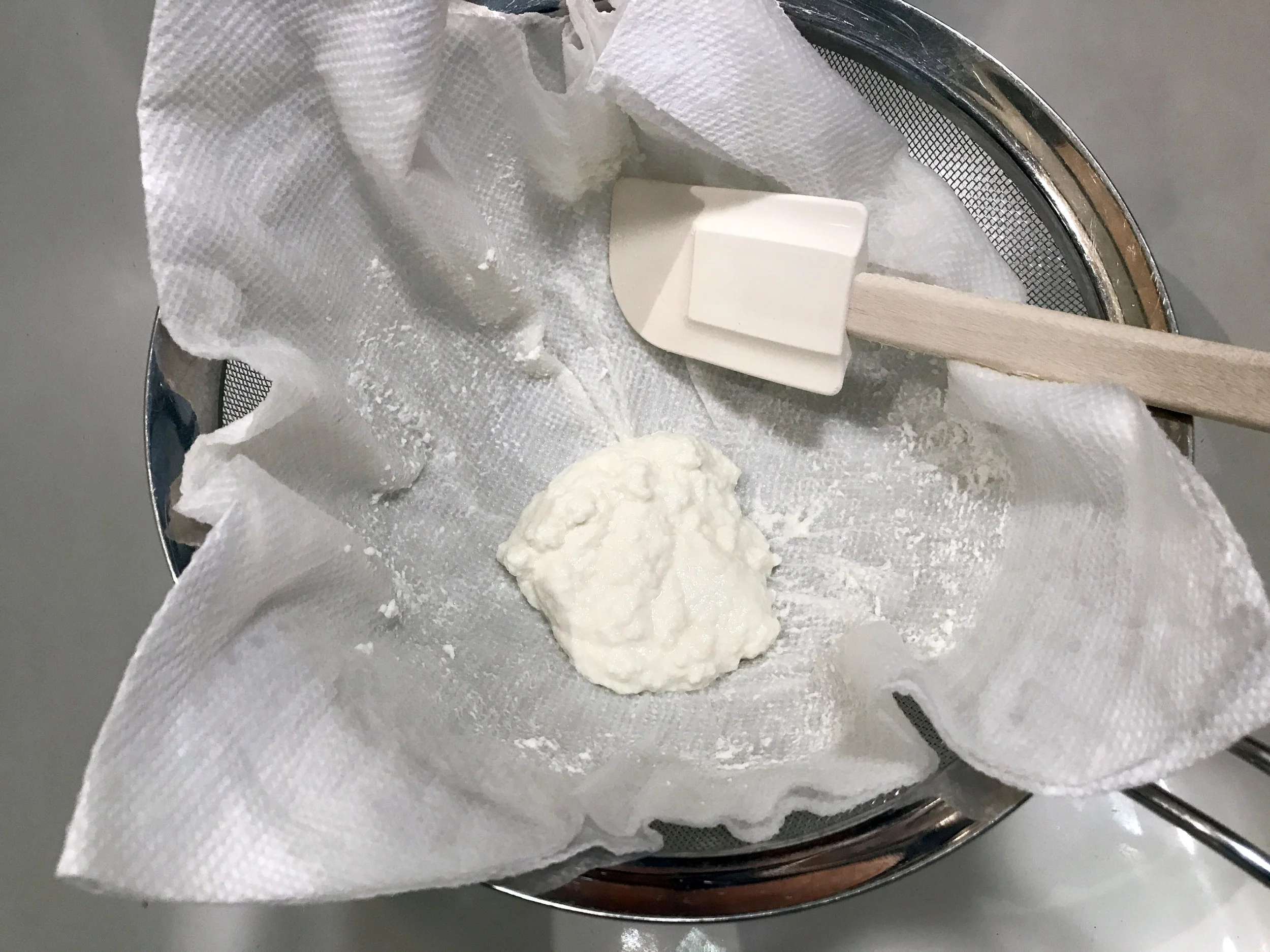 Ricotta draining in a cheesecloth-lined fine-mesh strainer set over a bowl on a kitchen counter