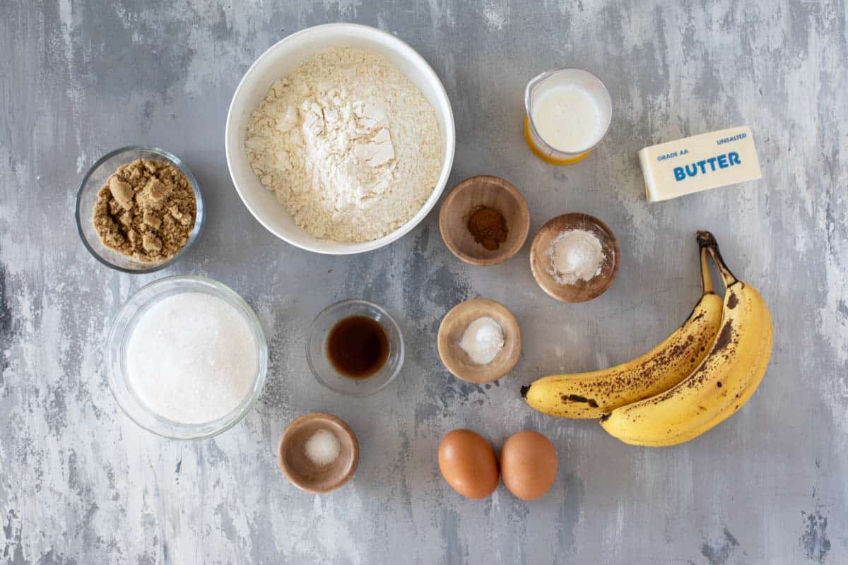 Ripe bananas, flour, sugar, eggs, butter, chocolate chips, and baking soda arranged on a clean kitchen counter with warm natural light