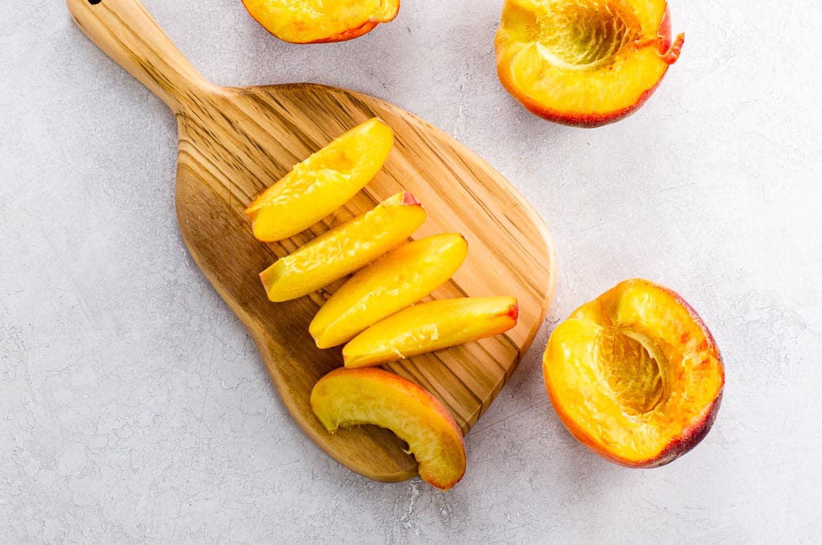 Ripe yellow peaches cut in half with pits removed, arranged on a cutting board with a small bowl of brown sugar and a stick of butter nearby on a bright kitchen counter