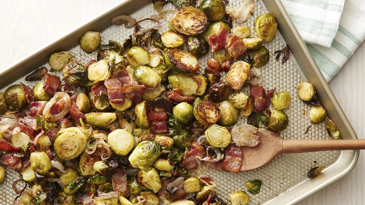 Roasted Brussels sprouts and bacon on a sheet pan being tossed with a spoon after roasting