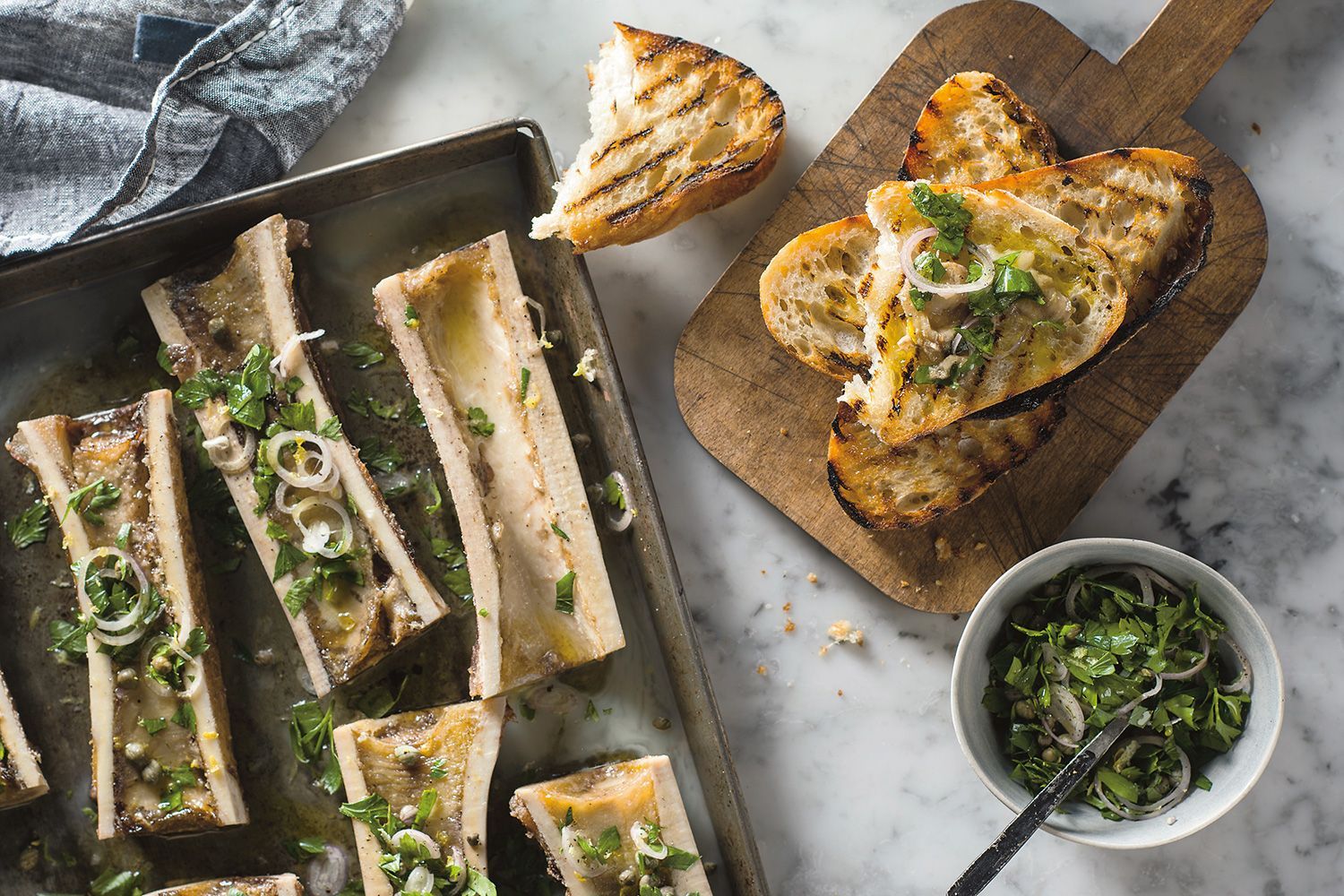 Roasted beef marrow bones on a baking sheet with bubbling marrow, served with crisp garlic toast and a small bowl of parsley salad on a warm kitchen table